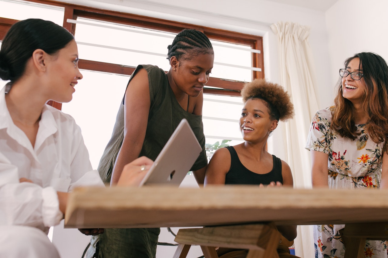 four women sitting by a desk in front of a computer, discussing