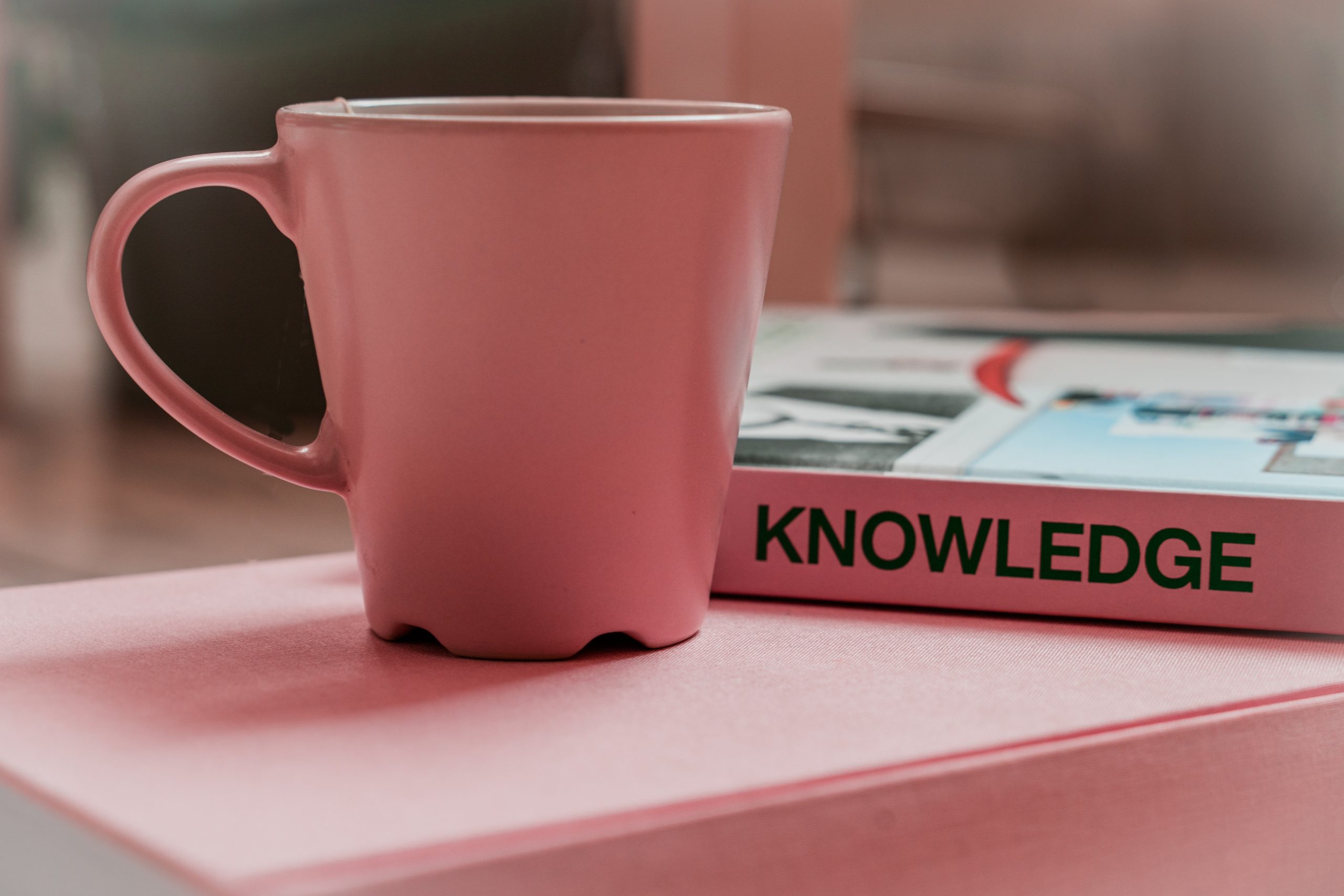 Pink cup on the pink table, behind there is a pink book titled Knowledge